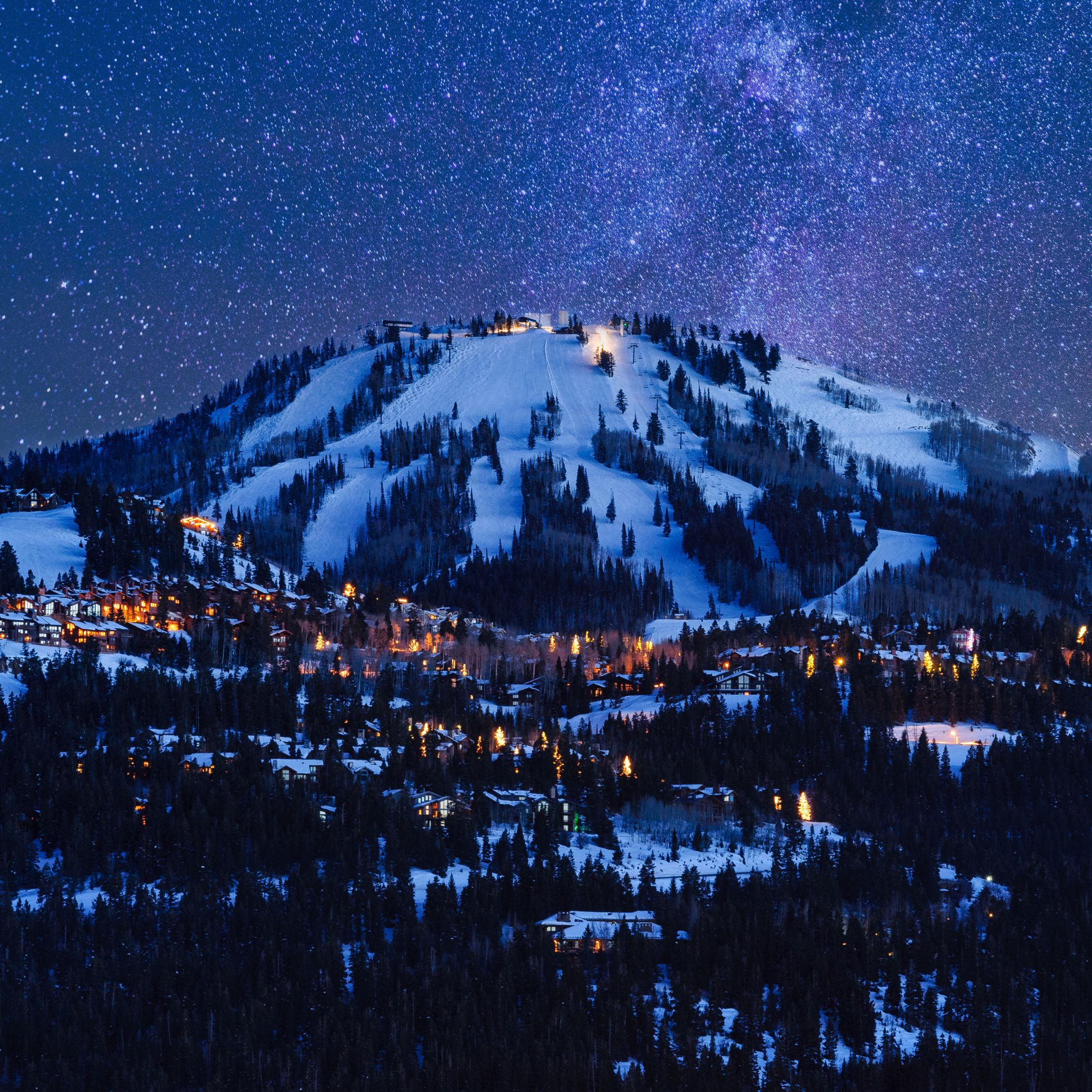 Nighttime image of the small town of Deer Valley at the foot of a massive mountain, illuminated under a beautiful starry sky with snow covering the ground, representing a perfect winter getaway 