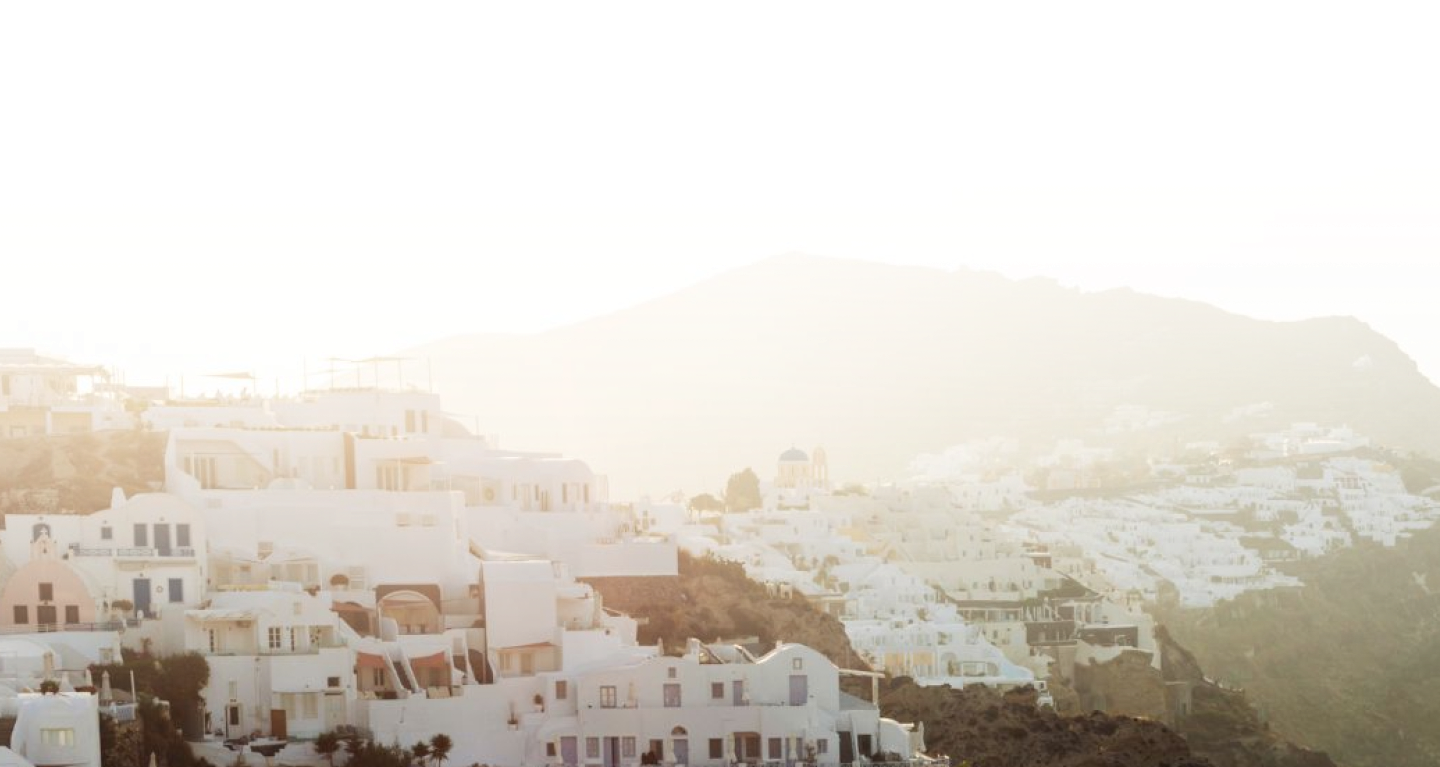 Iconic white Grecian buildings in a sunset cityscape, surrounded by green hills, captured from an aerial perspective